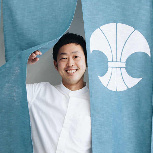 のれんブランド「中むら」CEO・中村 新さん。六本木とらやのれんも手掛けた若きプロデューサーが、自社の家紋入りのれんをめくって笑顔で登場