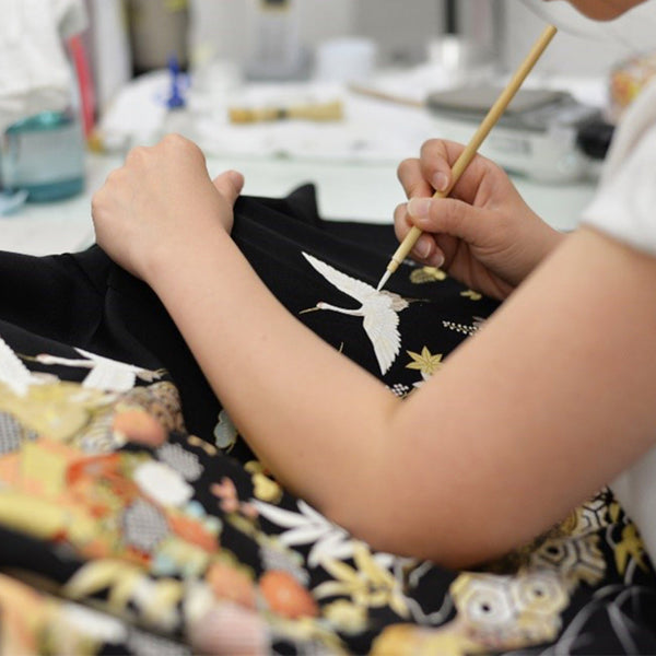 京都の着物アップサイクルブランド「季縁」の職人が、黒留袖の鶴の刺繍に細筆で仕上げを施す。着物を現代のクチュールドレスへと生まれ変わらせる制作の一場面