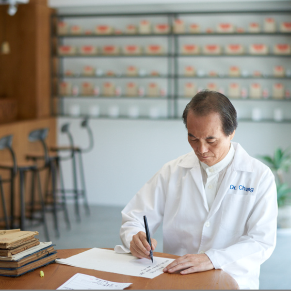 台北の漢方薬局「意一堂」院長・江 錦輝氏。白衣を纏い薬棚に囲まれた診療所で、40年の臨床経験をもとに漢方の処方を書き記す