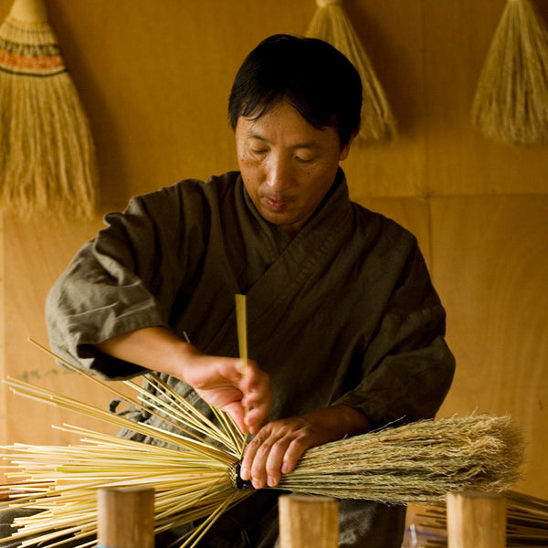 岩手県九戸村「高倉工芸」代表・高倉 清勝さん。作務衣姿で縮れたホウキモロコシの穂を束ねる。絹糸で編み上げる南部箒は20年使い続けられる逸品