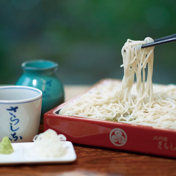 総本家更科堀井の更科そば。朱塗りの盆に盛られた純白の麺を箸で持ち上げた様子。「さらしな」と書かれた湯呑みと青磁の徳利を添えて